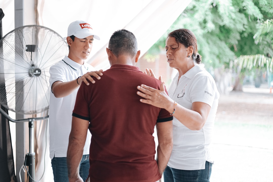 A man in a maroon shirt is comforted by a woman and a younger man at Health and Fitness Professionals, possibly related to workers comp physical therapy NJ.