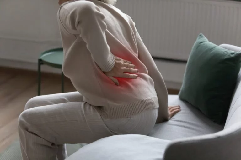 An older adult getting up from a sofa with a hand pressed to the hip and lower back, the sore area highlighted in red